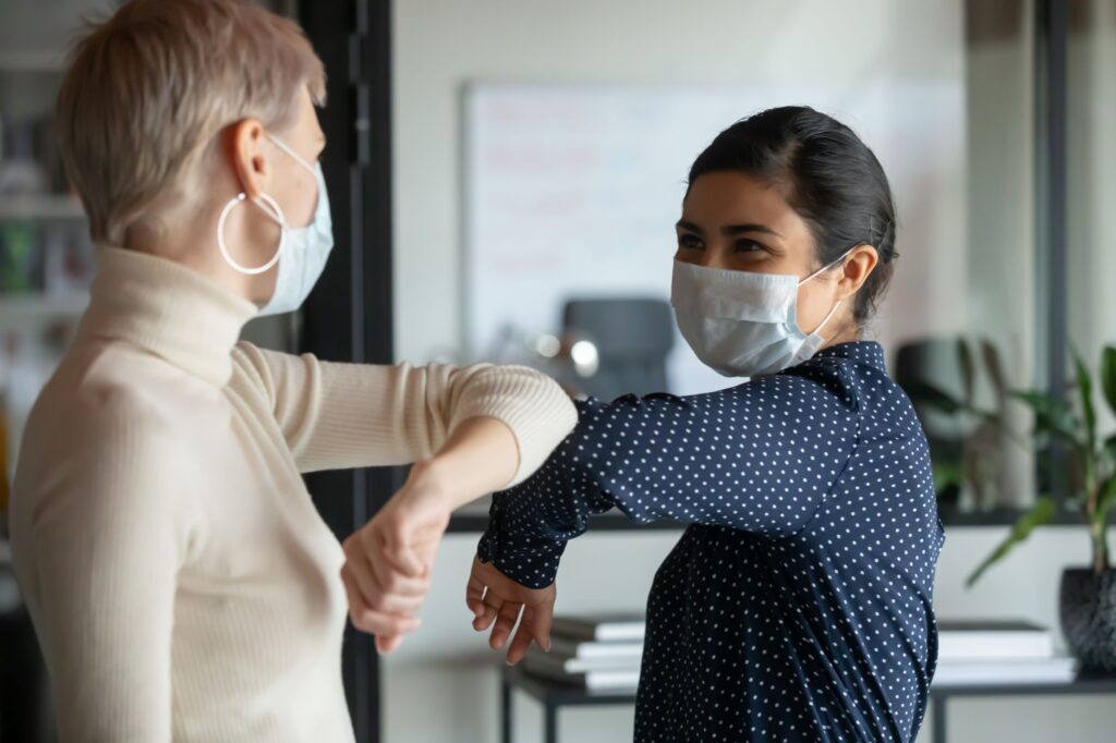 Two women wearing masks greet each other by bumping elbows to ensure health safety.