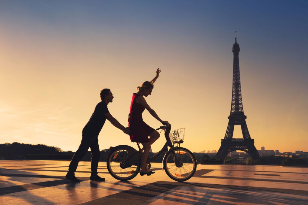 People enjoying a Paris sunset in front of the Eiffel Tower. One riding a bike while the other pushing for momentum.