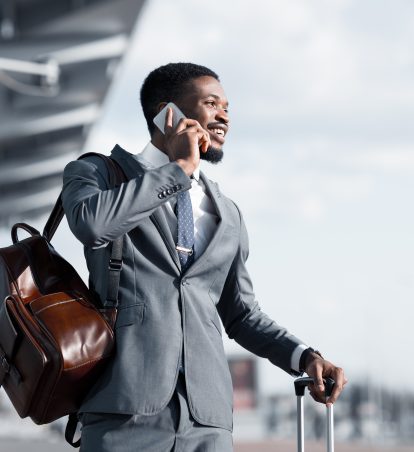 A business man standing at the airport, holding a suitcase in one hand and talking on his phone.