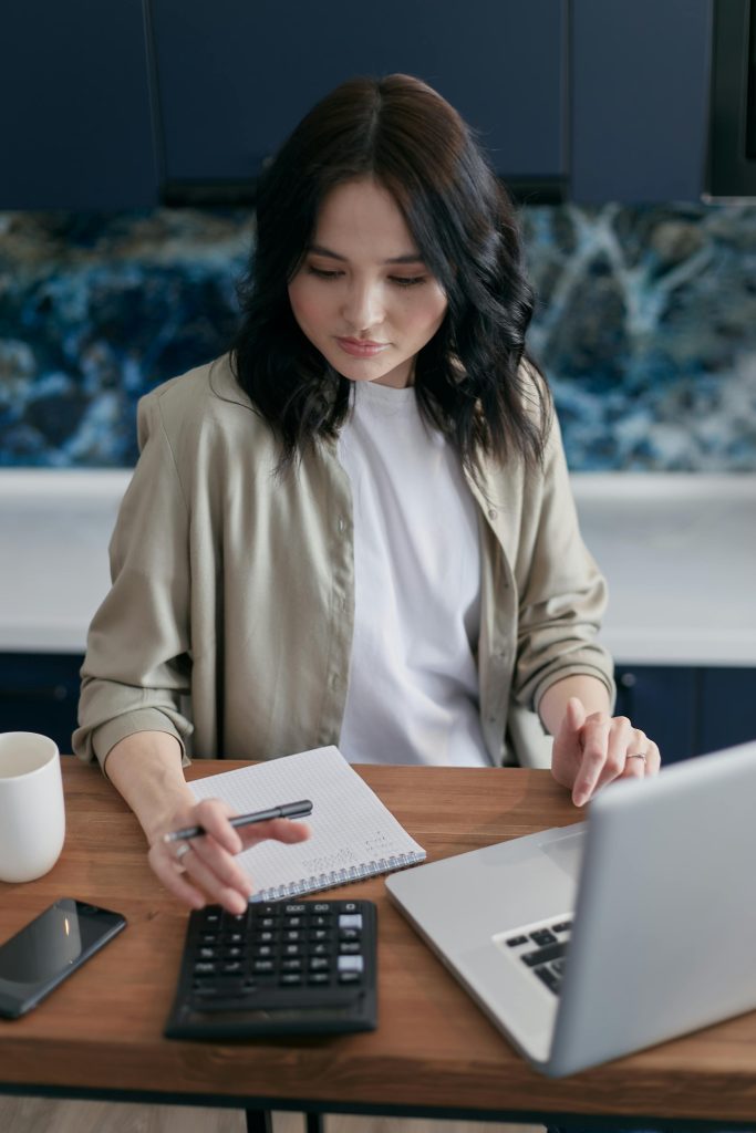 Woman Using a Calculator While Sitting In Front of a Laptop. 